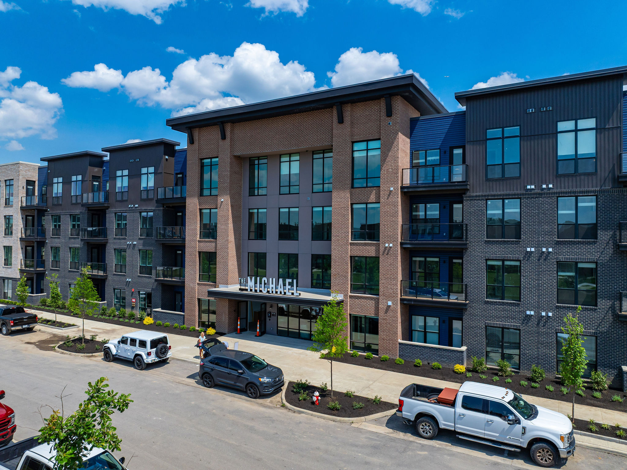 Thrive Companies, A modern four-story apartment building with brick and dark panel exterior, large windows, and several cars parked in front under a partly cloudy sky awaits the excitement of the Michael Grand Opening. Apartments in Columbus Ohio