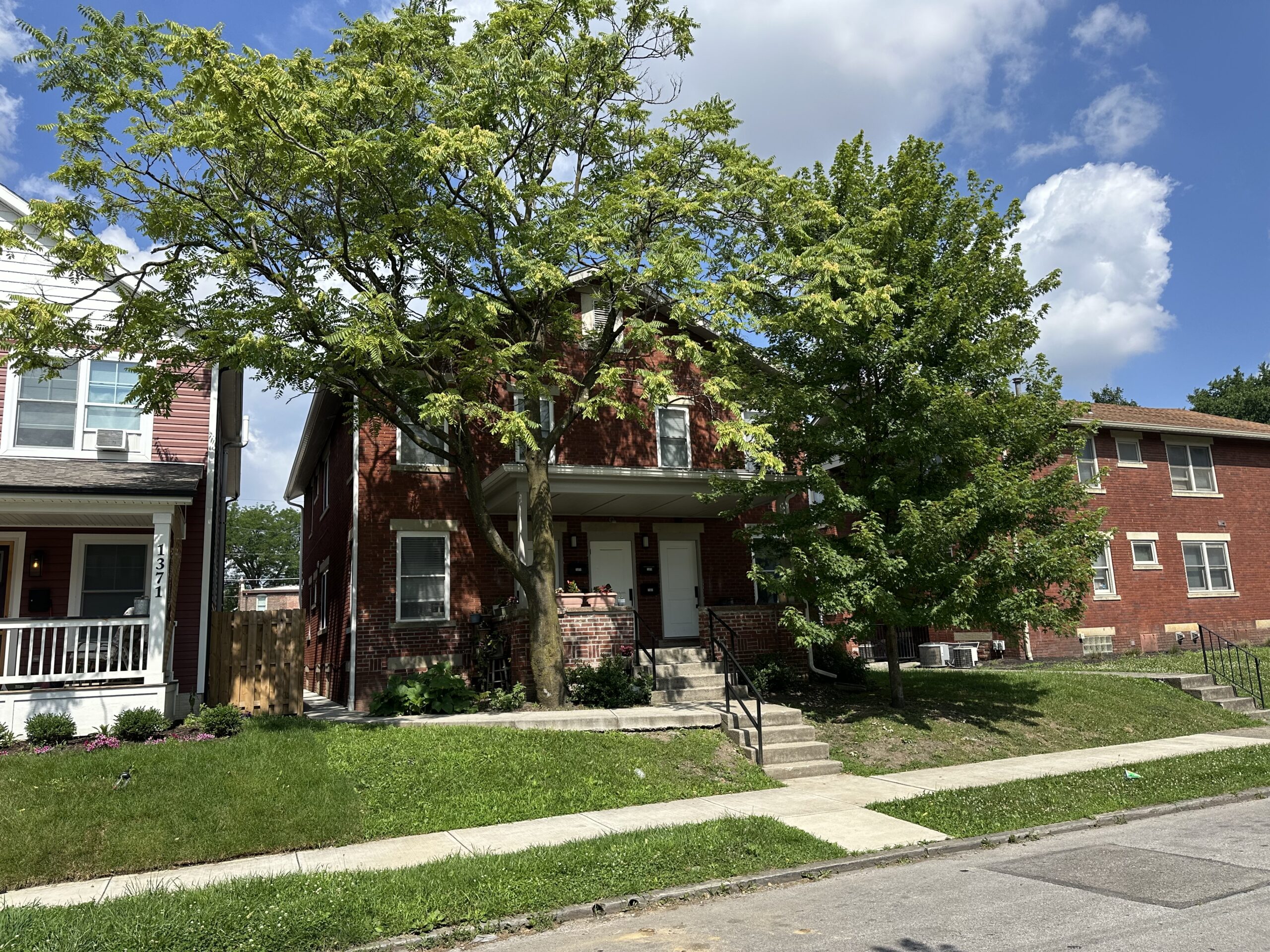 Thrive Companies, Two-story red brick house with a covered porch, shaded by large trees, sits next to a sidewalk on a sunny day. Neighboring buildings are visible on both sides. Apartments in Columbus Ohio