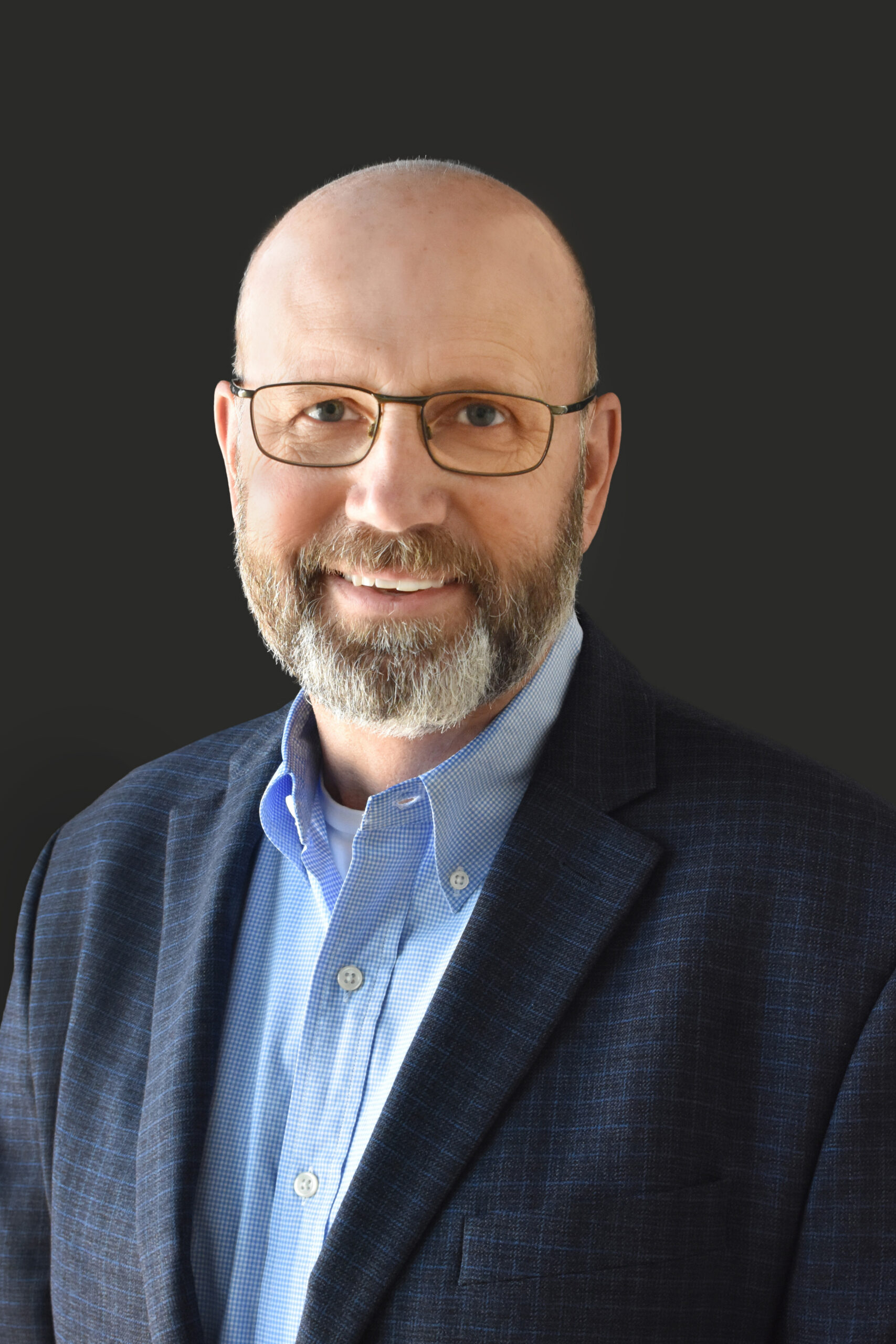 Thrive Companies, A member of Our Team, this middle-aged man with glasses, a bald head, and a gray beard, wearing a blue dress shirt and dark blazer, poses confidently in front of a plain dark background. Apartments in Columbus Ohio