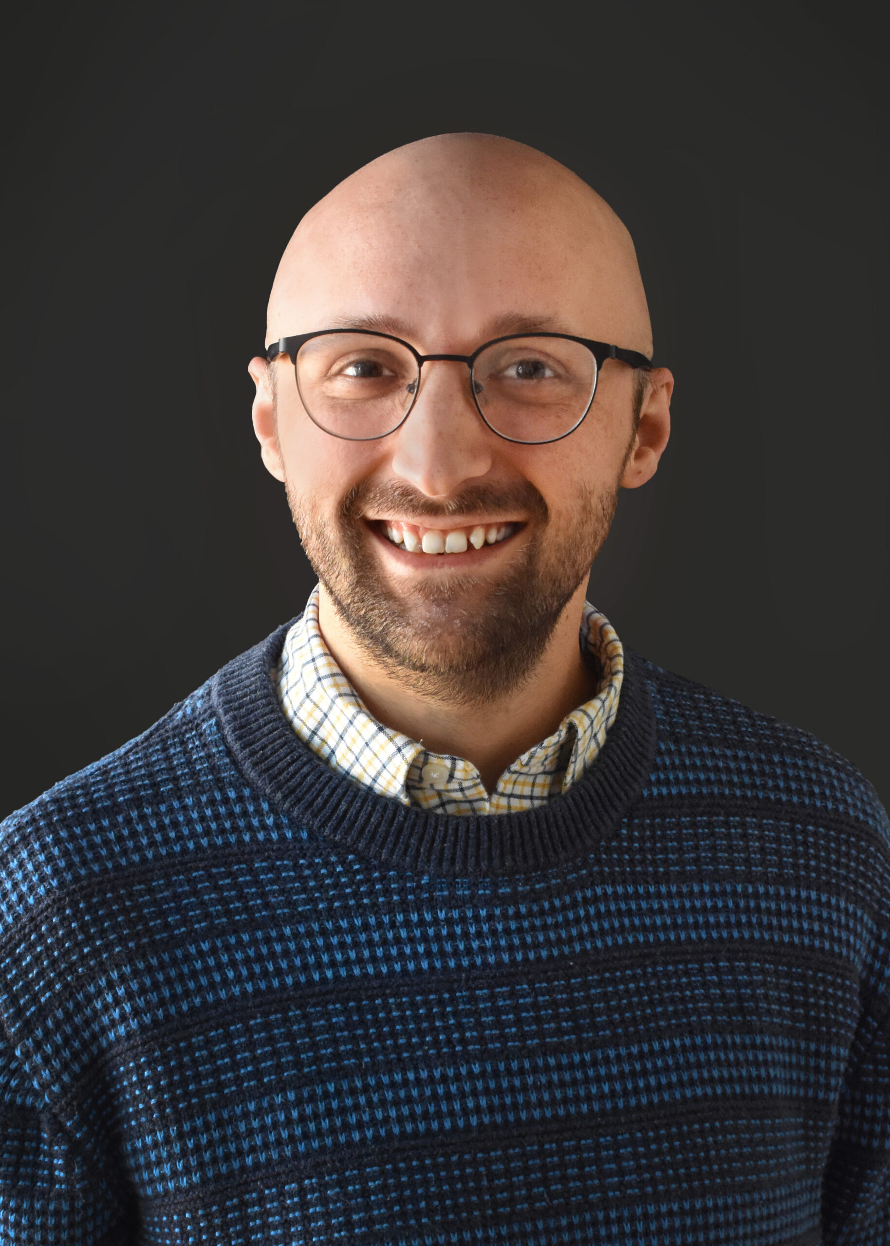 Thrive Companies, A smiling member of Our Team with glasses and a bald head, wearing a blue patterned sweater over a checkered shirt, poses against a plain dark background. Apartments in Columbus Ohio
