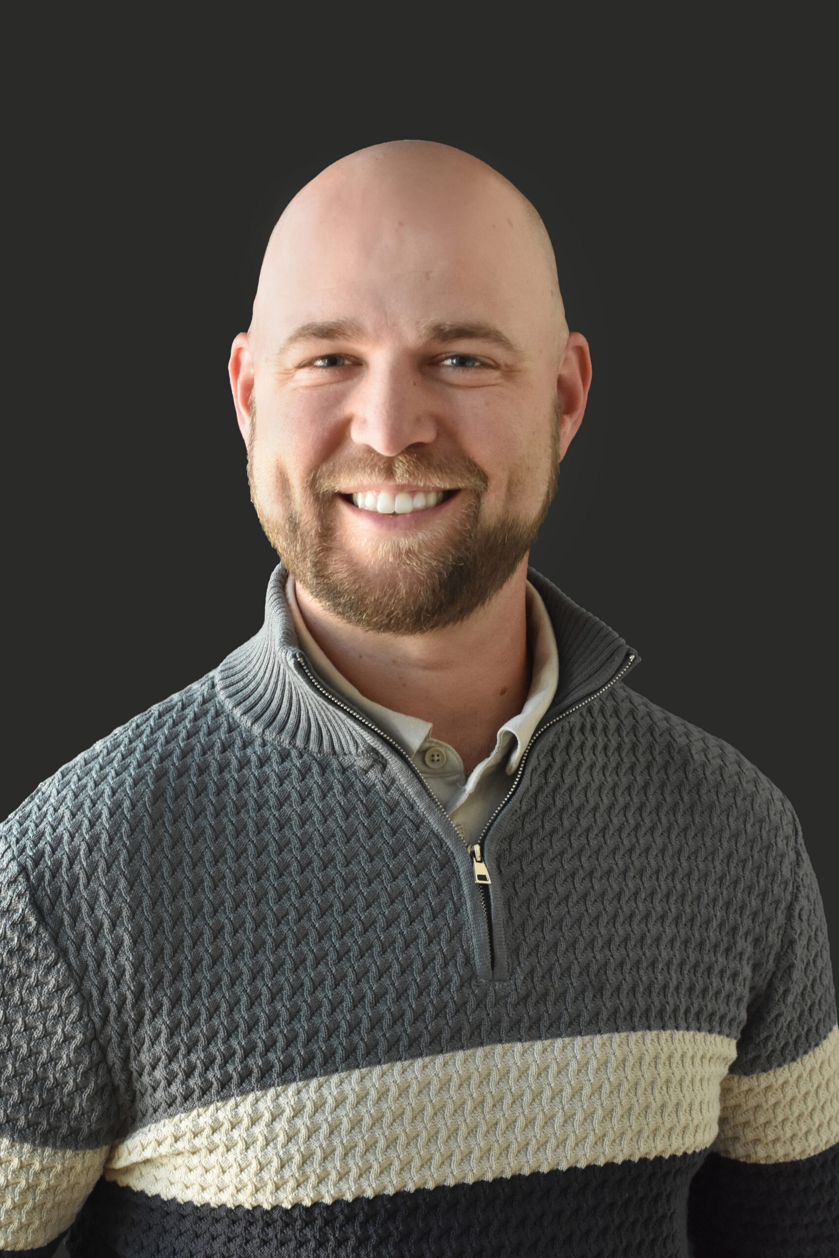 Thrive Companies, A member of Our Team, a bald man with a beard smiles at the camera, wearing a gray patterned pullover sweater with a light stripe and collared shirt underneath, in front of a plain dark background. Apartments in Columbus Ohio