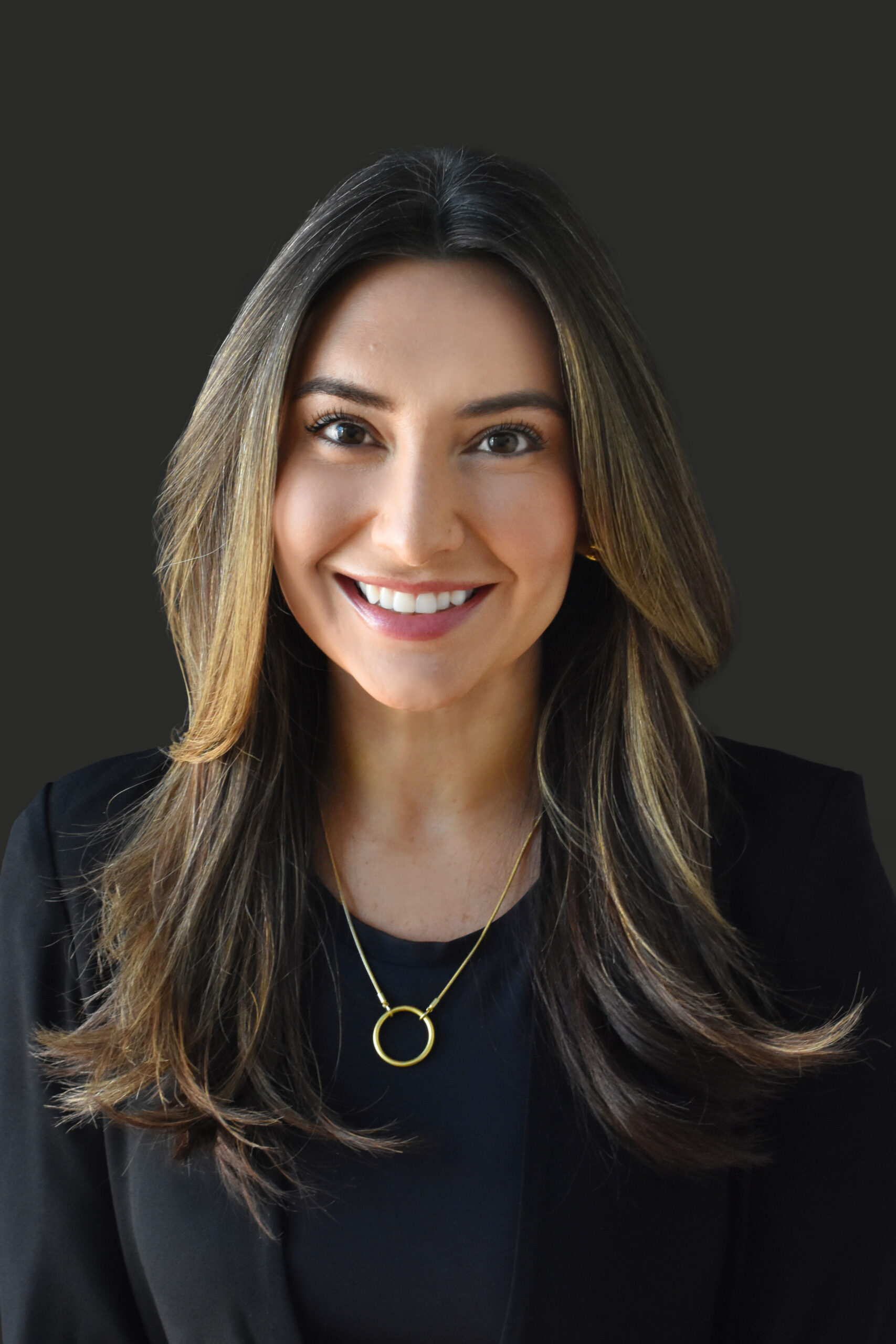 Thrive Companies, Our Team member, a woman with long brown hair, wears a black blazer, black top, and gold necklace, smiling confidently in front of a plain dark background. Apartments in Columbus Ohio