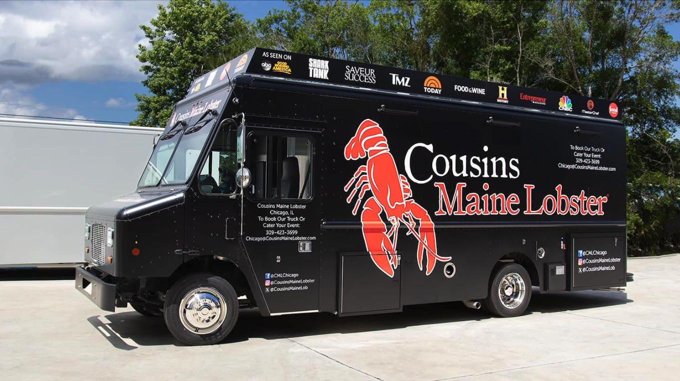 Thrive Companies, A black food truck with the Cousins Maine Lobster logo and a large red lobster graphic is parked on a concrete lot with trees in the background, ready to serve up delicious seafood favorites this Friday. Apartments in Columbus Ohio