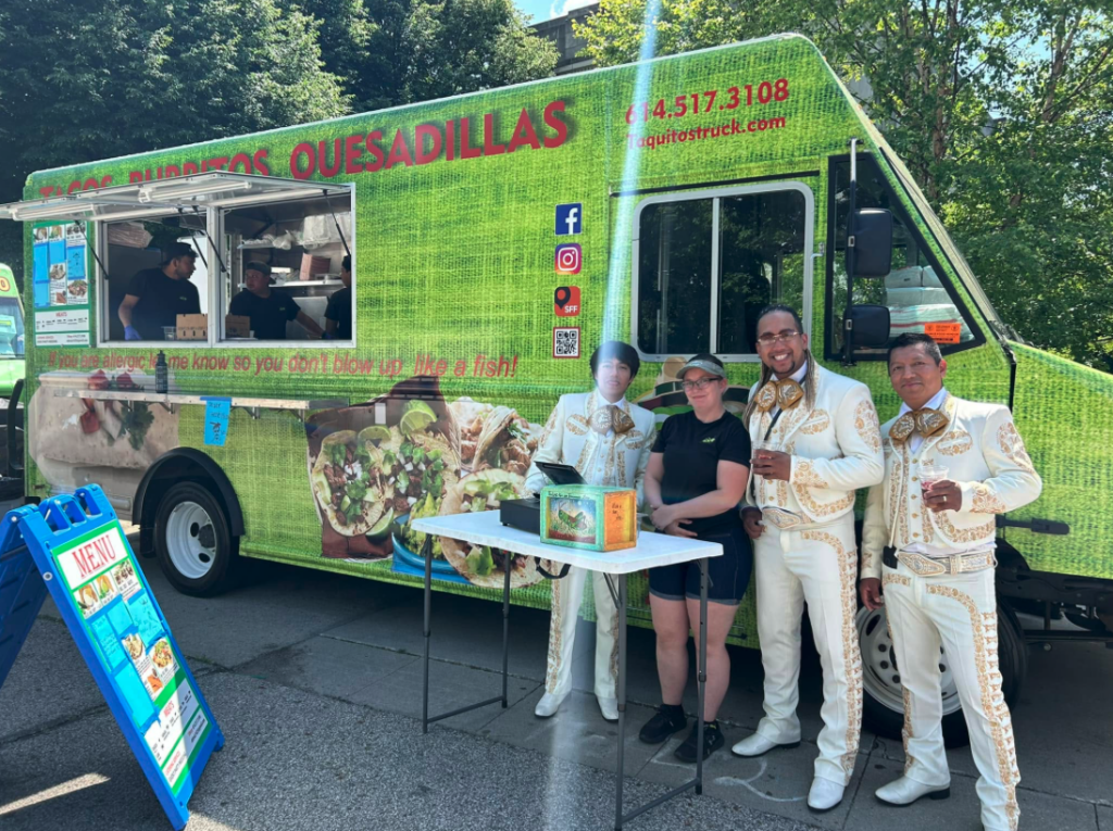Thrive Companies, Four people pose in front of a green food truck at Food Truck Friday; three wear matching mariachi outfits and one wears casual clothes. A small table with items, possibly including taquitos, sits in front of them. Apartments in Columbus Ohio