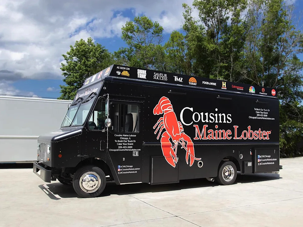 Thrive Companies, The Cousins Maine Food Truck features a large red lobster graphic and serves fresh Maine lobster; it’s parked outdoors on a sunny day with trees and a white truck nearby. Apartments in Columbus Ohio